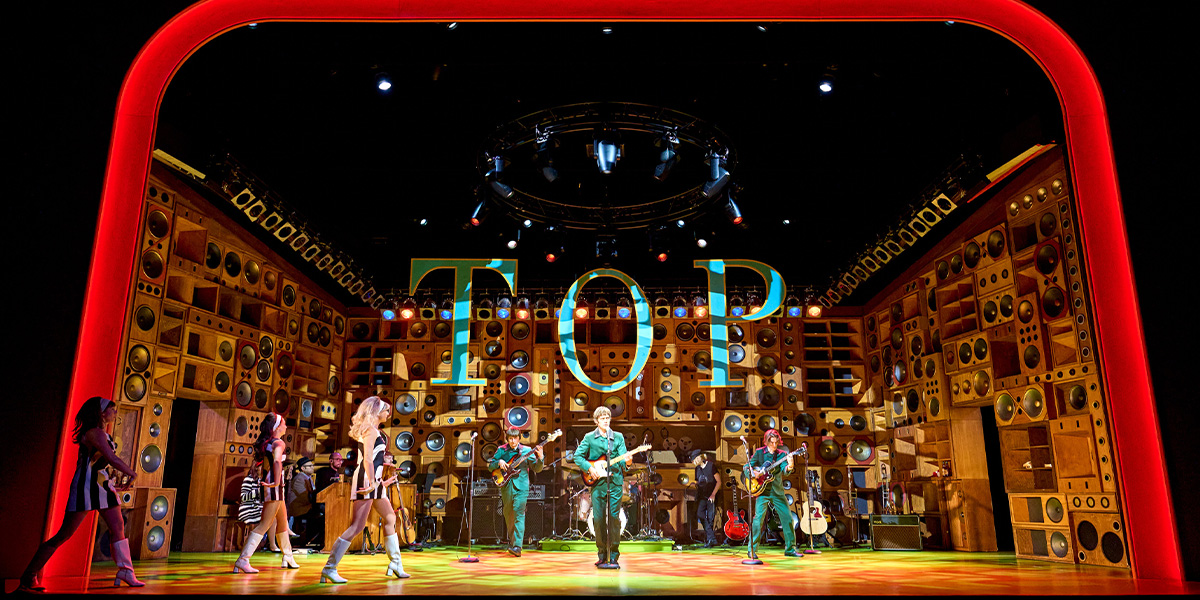 A group of performers in vibrant costumes are singing on stage, with a bright backdrop and stage lights.