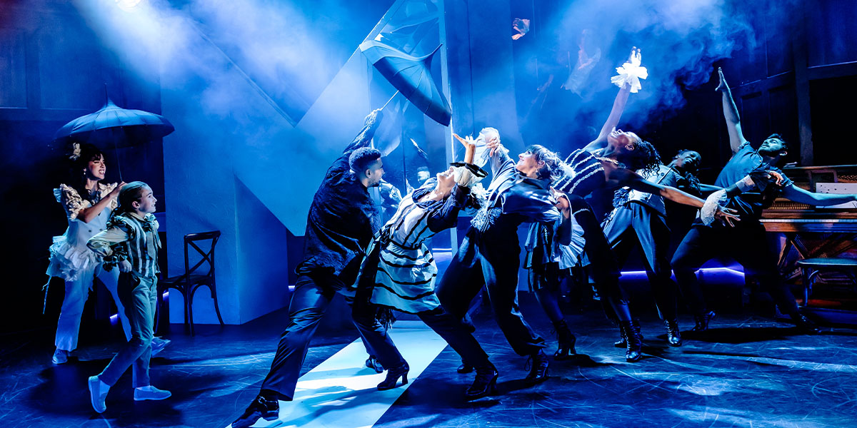 A dynamic scene from a theatrical production with performers in various costumes, some holding umbrellas, dancing energetically on a dramatically lit stage.