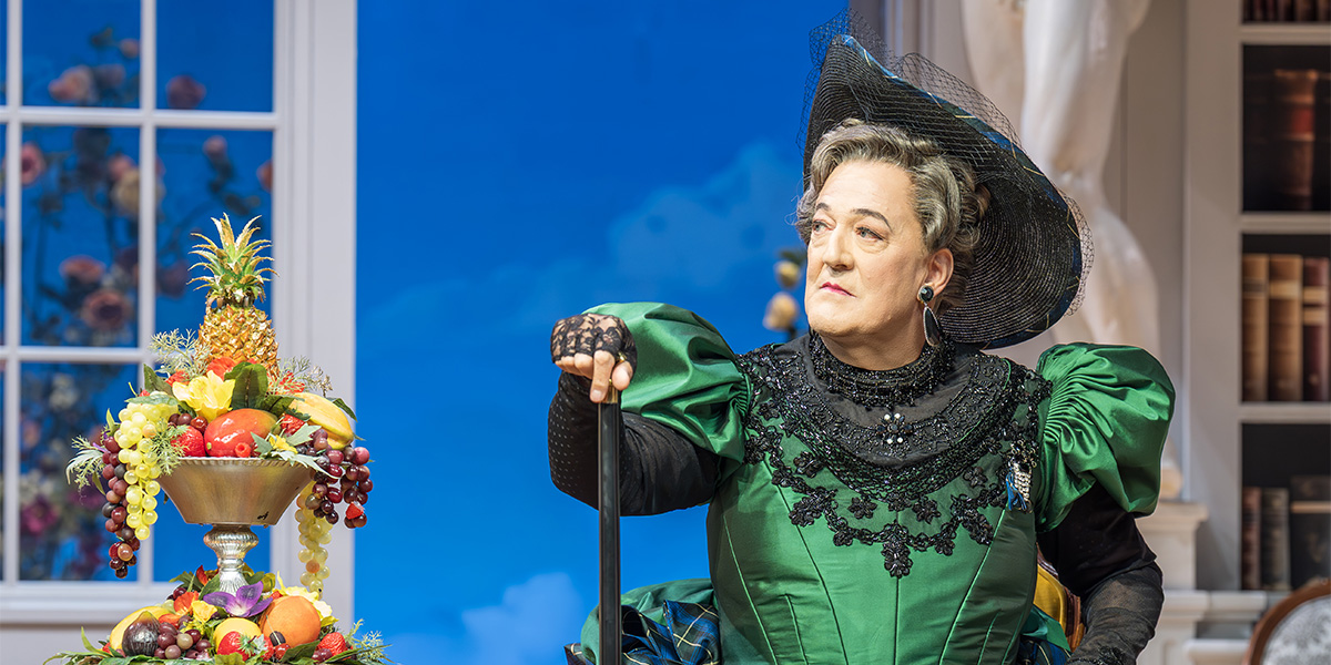 Stephen Fry, dressed in formal attire, gestures expressively on stage, surrounded by theatrical set elements.