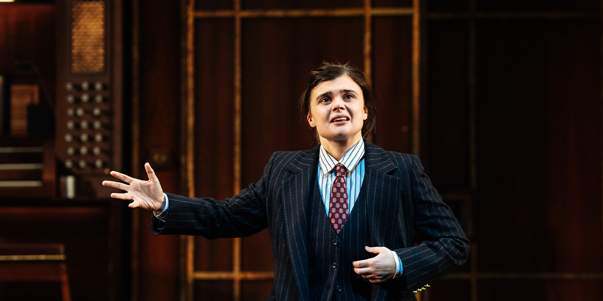 A performer in a pinstripe suit gestures expressively, with a wooden backdrop and stage lighting.