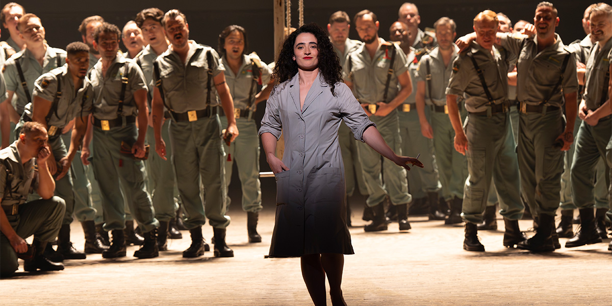 A diverse chorus in vibrant costumes performs passionately on stage, with dramatic lighting enhancing the scene.