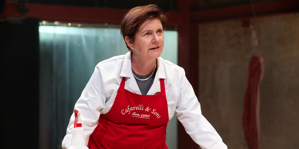 Jackie Clune, wearing a red apron, engages with the audience in a theatrical setting.