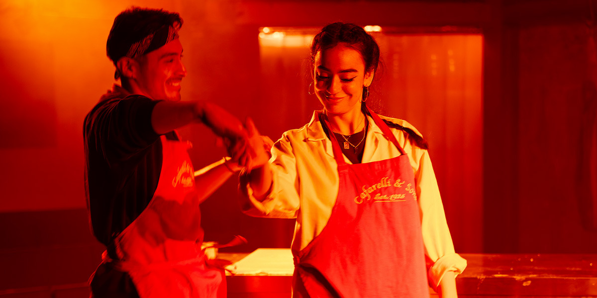 Marcello Cruz and Mithra Malek share a joyful moment, both wearing aprons, in warm red lighting.