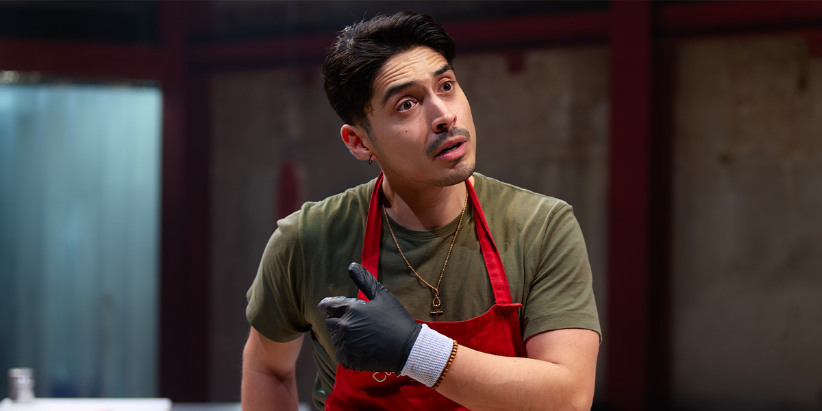 Marcello Cruz, wearing a red apron and black gloves, gestures while speaking in a rustic setting.