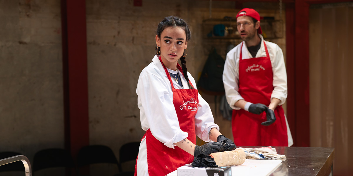 Mithra Malek and Eugene McCoy, both in red aprons, prepare food in a rustic kitchen setting.