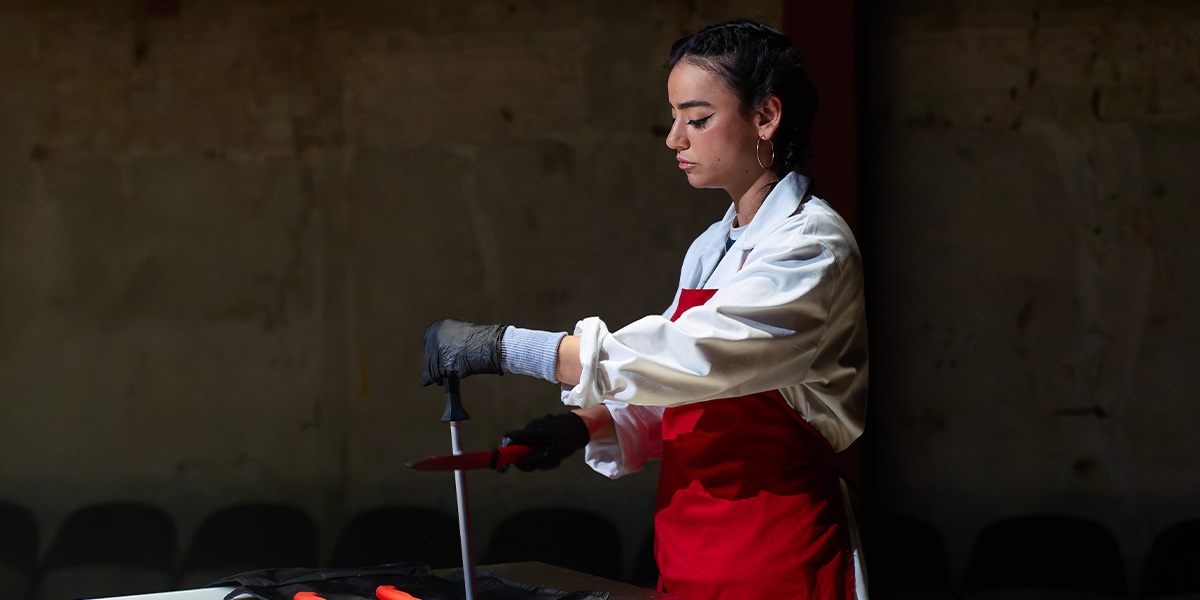 Mithra Malek, wearing a red apron and gloves, skillfully handles a knife at a table.