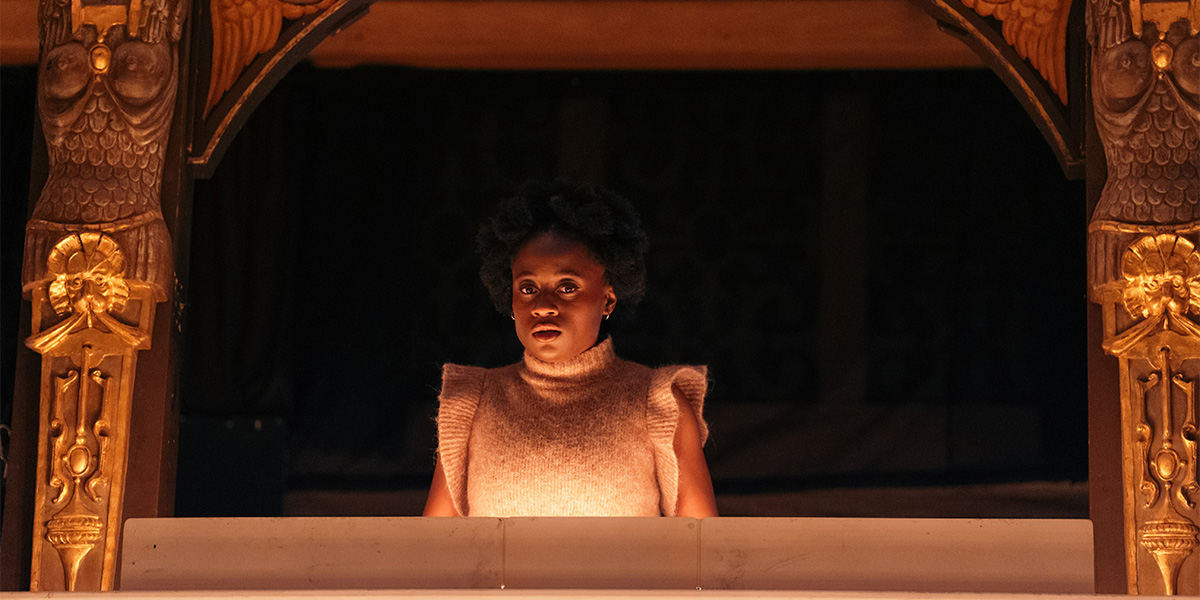 Tara Tijani as Helena, wearing a flowing green dress, stands centre stage at Shakespeare's Globe.