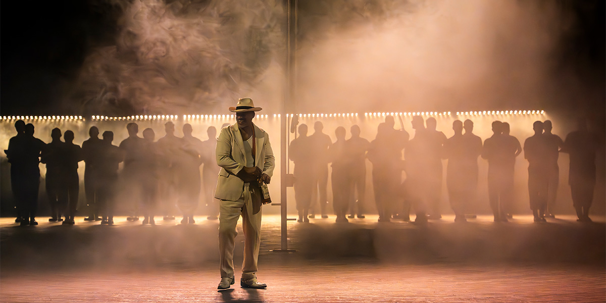 A diverse cast in elaborate costumes poses dramatically on stage, with vibrant colours and theatrical lighting.