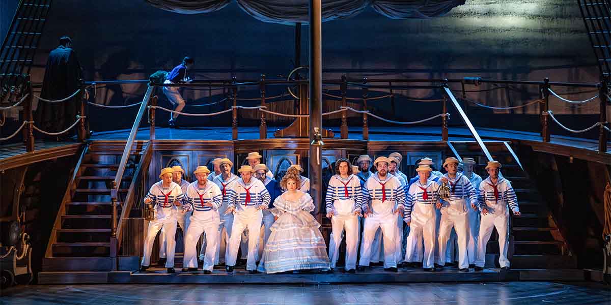 The cast of ENO's HMS Pinafore, dressed in sailor outfits, performs on a ship-themed stage.