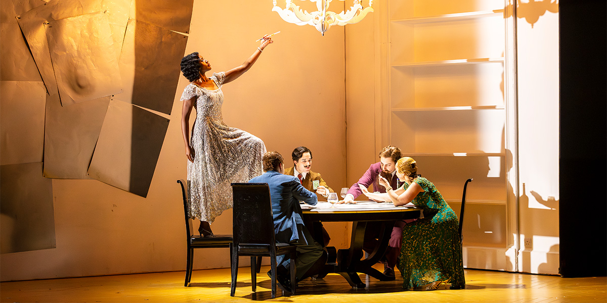 The cast of ENO's Partenope 2025 poses together, wearing elaborate costumes in vibrant colours against a dramatic backdrop.