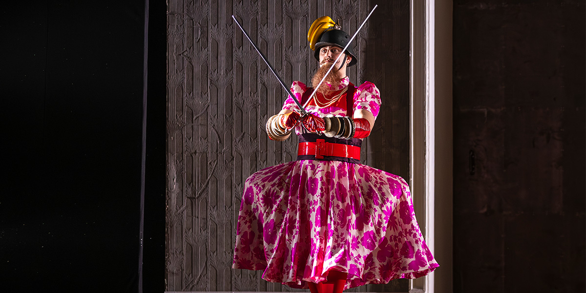 William Thomas as Partenope, wearing a vibrant costume, poses confidently against a dramatic stage backdrop.