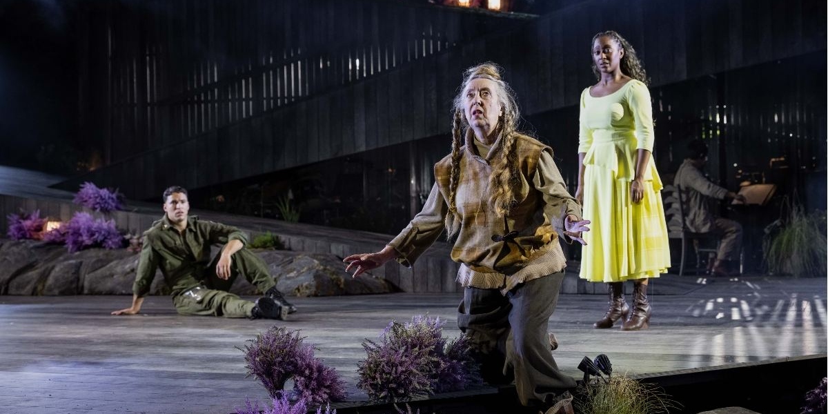 Three actors perform onstage: one kneels with dramatic expression, another sits on the ground in fatigues, and a third stands in a yellow dress. The stage is set with rocks, plants, and dim lighting.