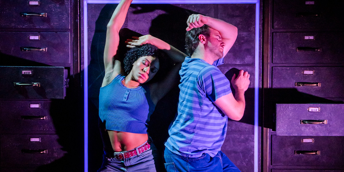 A group of four performers in vibrant costumes interact on stage, surrounded by dramatic lighting and set design.
