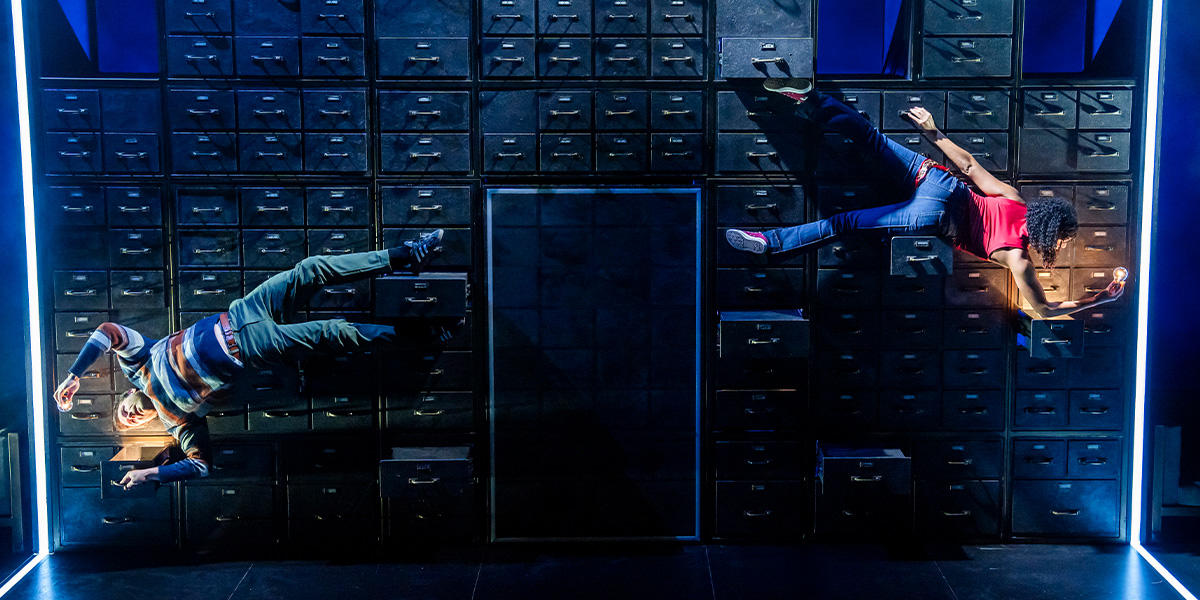 Four performers in vibrant costumes interact energetically on stage, surrounded by dramatic lighting and abstract set design.