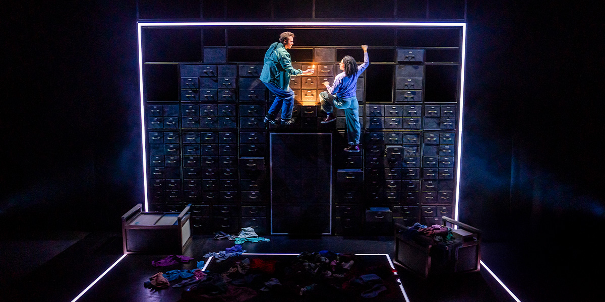 A group of four performers in vibrant costumes interact energetically on stage, with dramatic lighting and a textured backdrop.