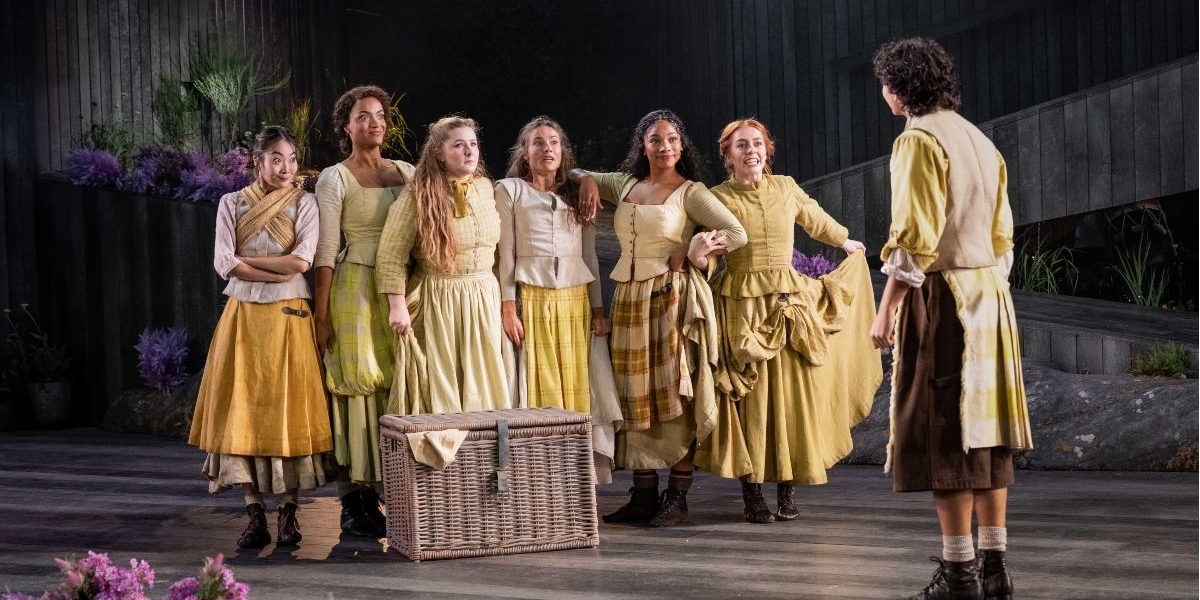 Six women in period costumes stand together on stage, smiling and facing a person with short dark hair in brown knickers and a yellow shirt. There is a wicker basket in front of the group and purple flowers in the background.