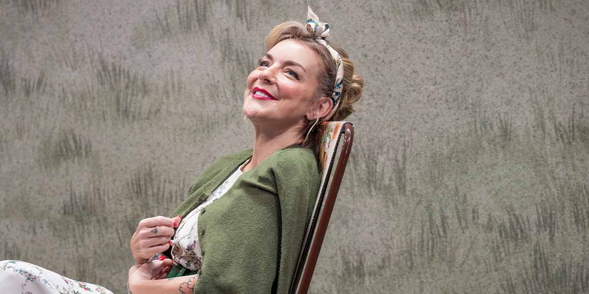 Sheridan Smith smiles while seated, wearing a floral dress and green cardigan, against a textured background.