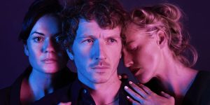 A dimly lit theatre stage with two actors in formal attire, engaged in an intense conversation.