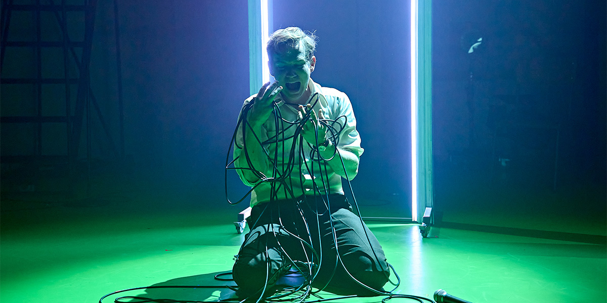 Jack Holden stands confidently in a modern costume, surrounded by dramatic lighting and a textured backdrop.