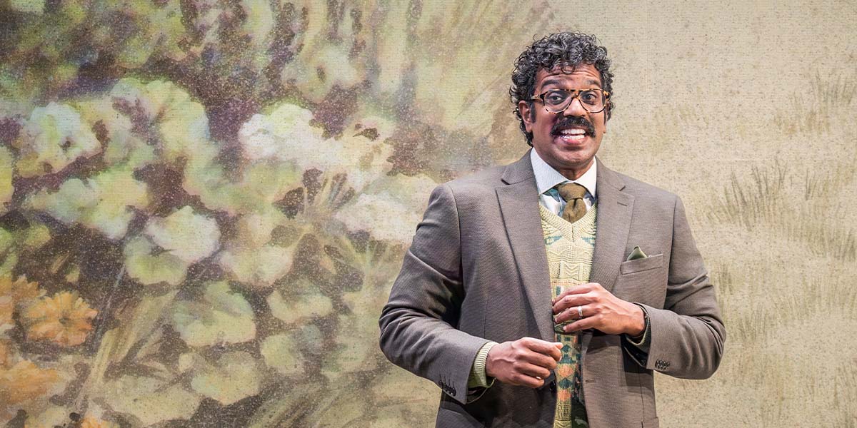 Romesh Ranganathan as Bill, sitting on a chair, wearing a blue shirt, with a neutral background.