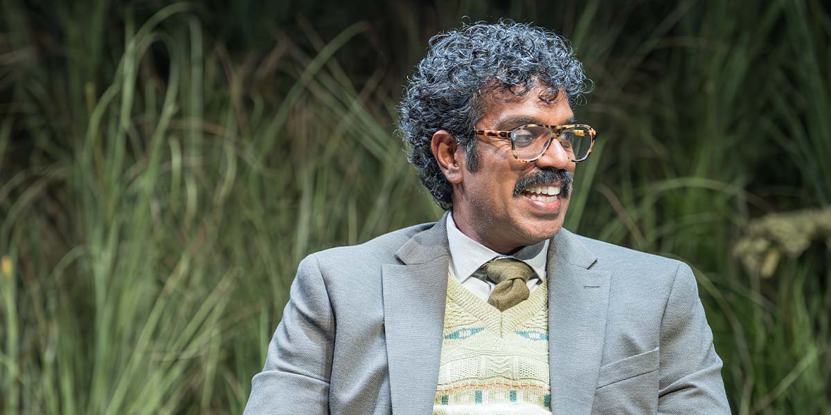 Romesh Ranganathan as Bill, seated on a chair, wearing a grey suit, with a thoughtful expression.