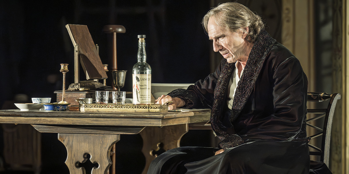 Ralph Fiennes as Henry Irving, dressed in period costume, gestures expressively against a dark, textured background.