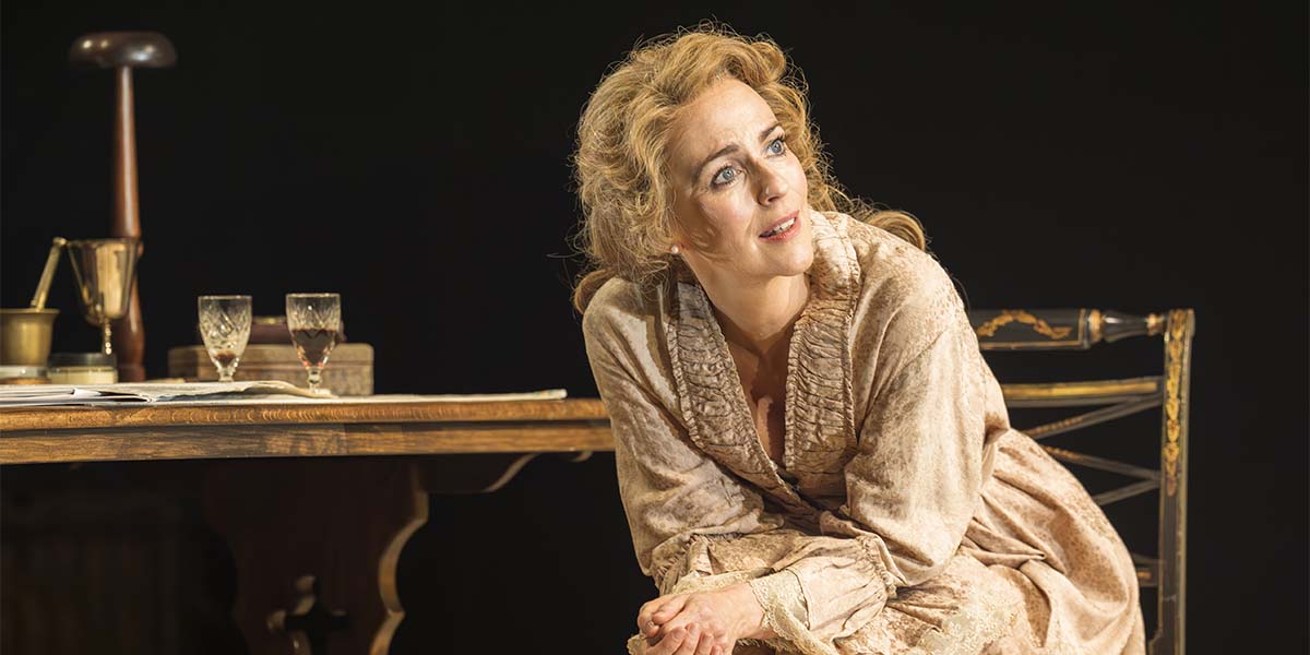 Miranda Raison, portraying Ellen Terry, gazes thoughtfully while seated beside a wooden table.