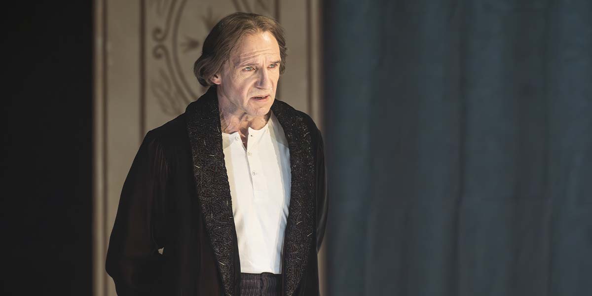 Ralph Fiennes, as Henry Irving, stands in a dark robe, looking serious against a softly lit backdrop.