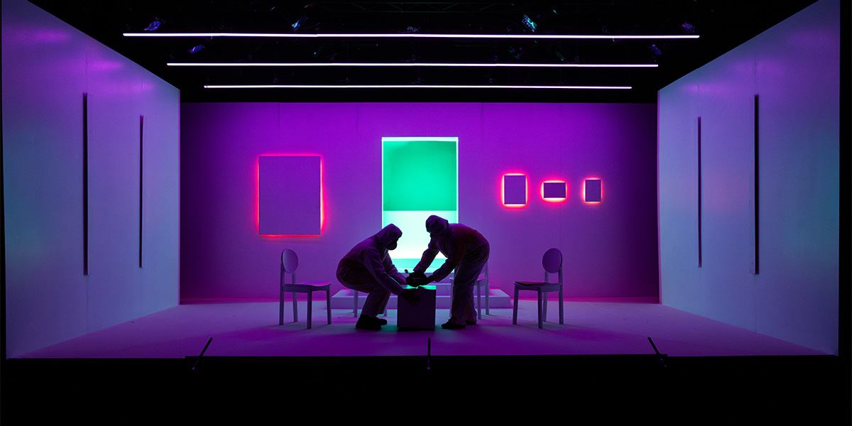 Two figures in protective suits interact with a box on a brightly lit, purple stage.