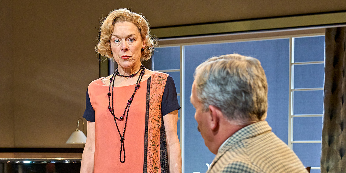 Janie Dee as Julia Steroll and Christopher Hollis as Bill Banbury pose together, smiling, in theatrical costumes.