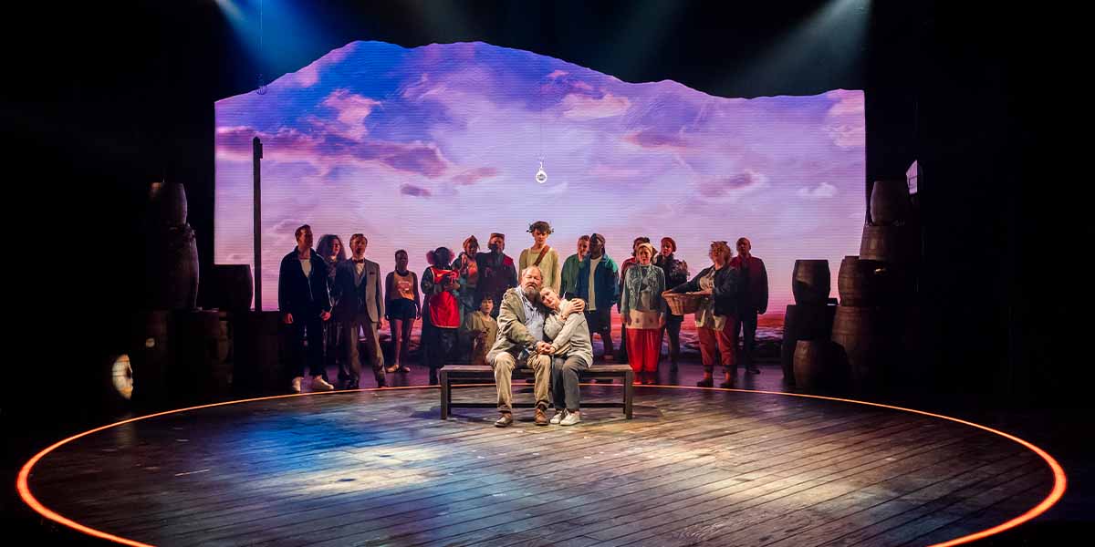 Mark Addy and Jenna Russell sit on a bench, surrounded by a cast against a scenic backdrop.