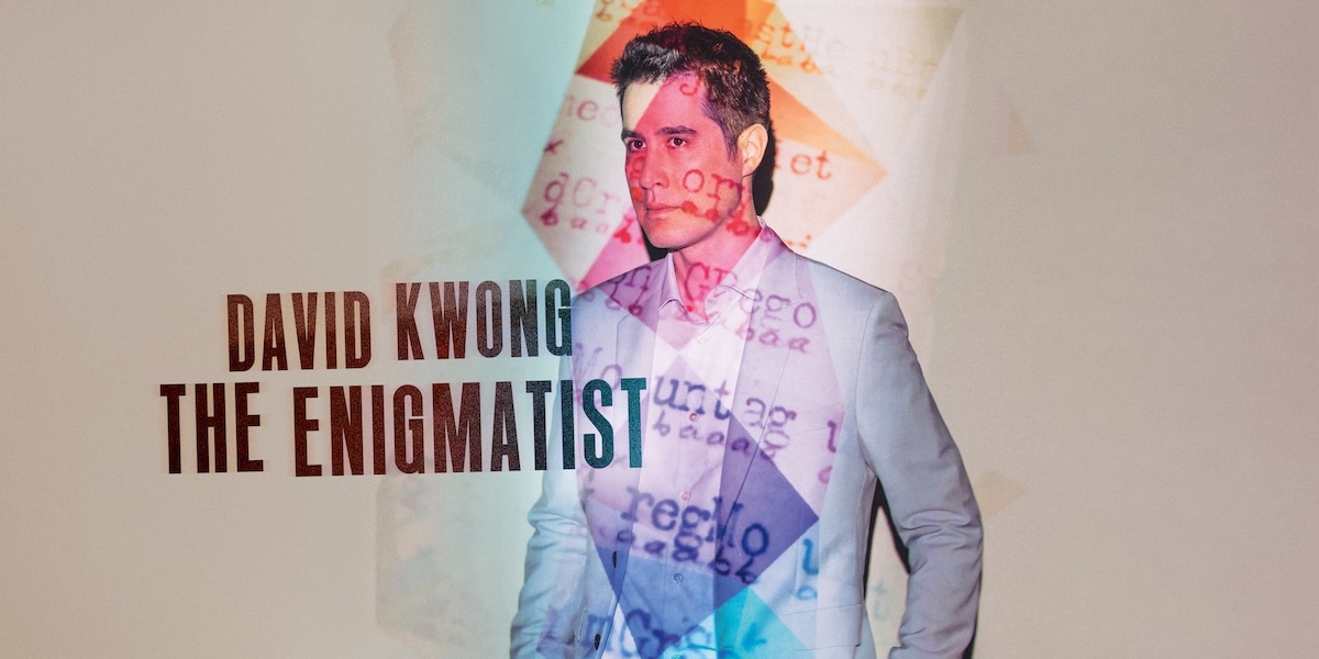 A mysterious figure in a dark suit stands in front of a large, intricate puzzle backdrop.