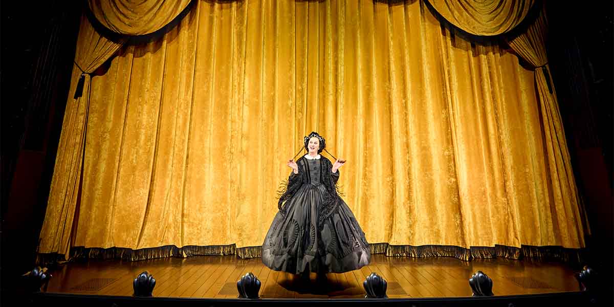 Jinkx Monsoon, dressed in a black gown, performs on stage before a golden curtain backdrop.