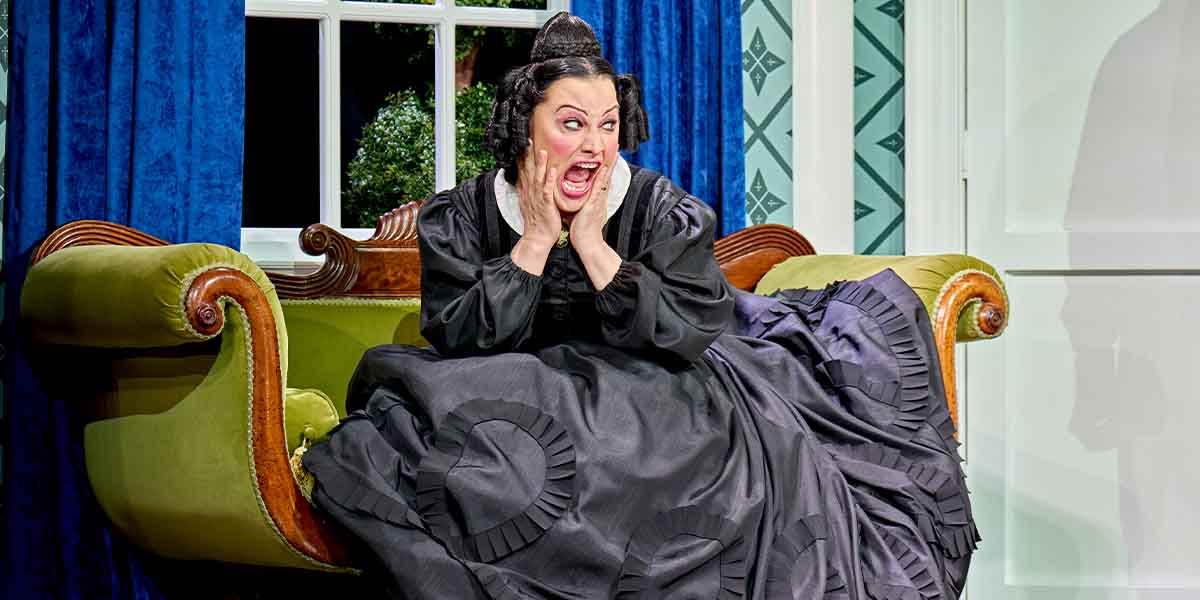 Jinkx Monsoon as Mary Todd Lincoln, seated on a green sofa, expressing shock in a black dress.