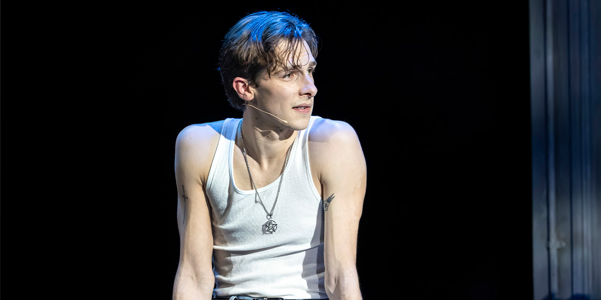 Killian Thomas Lefevre sits on stage, wearing a white tank top, with dramatic lighting highlighting his expression.