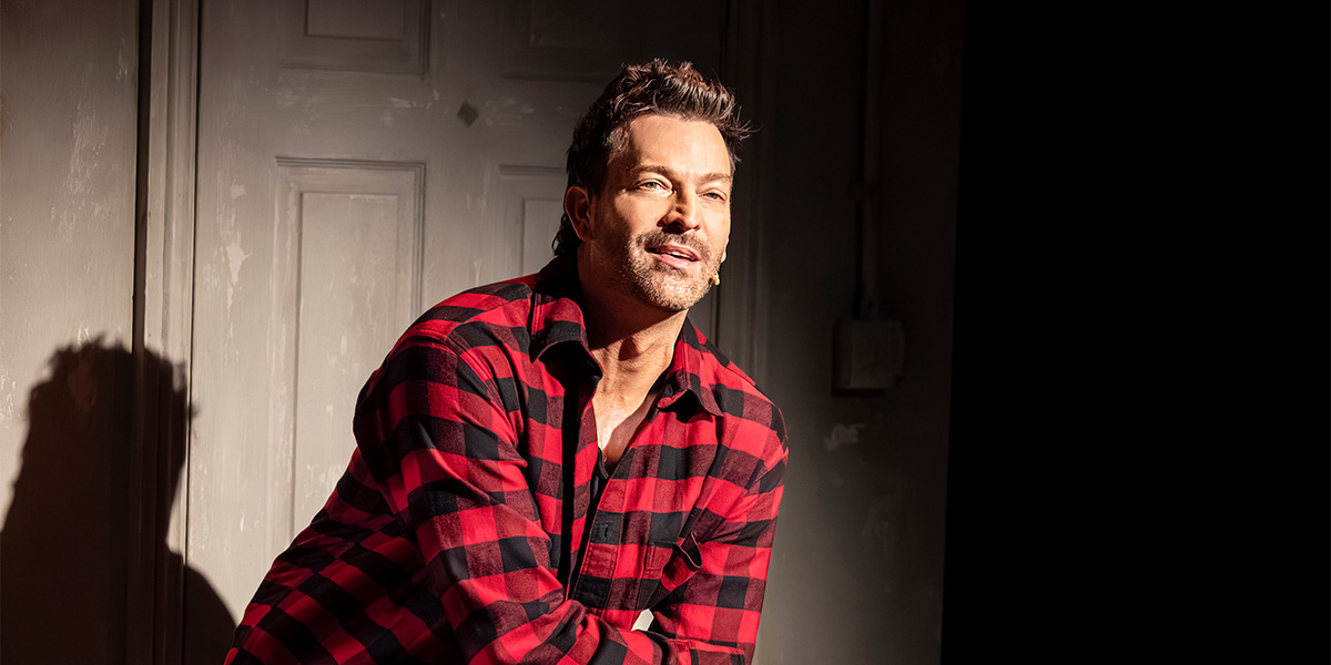 Levi Kreis smiles while wearing a red and black checkered shirt, posed against a grey backdrop.
