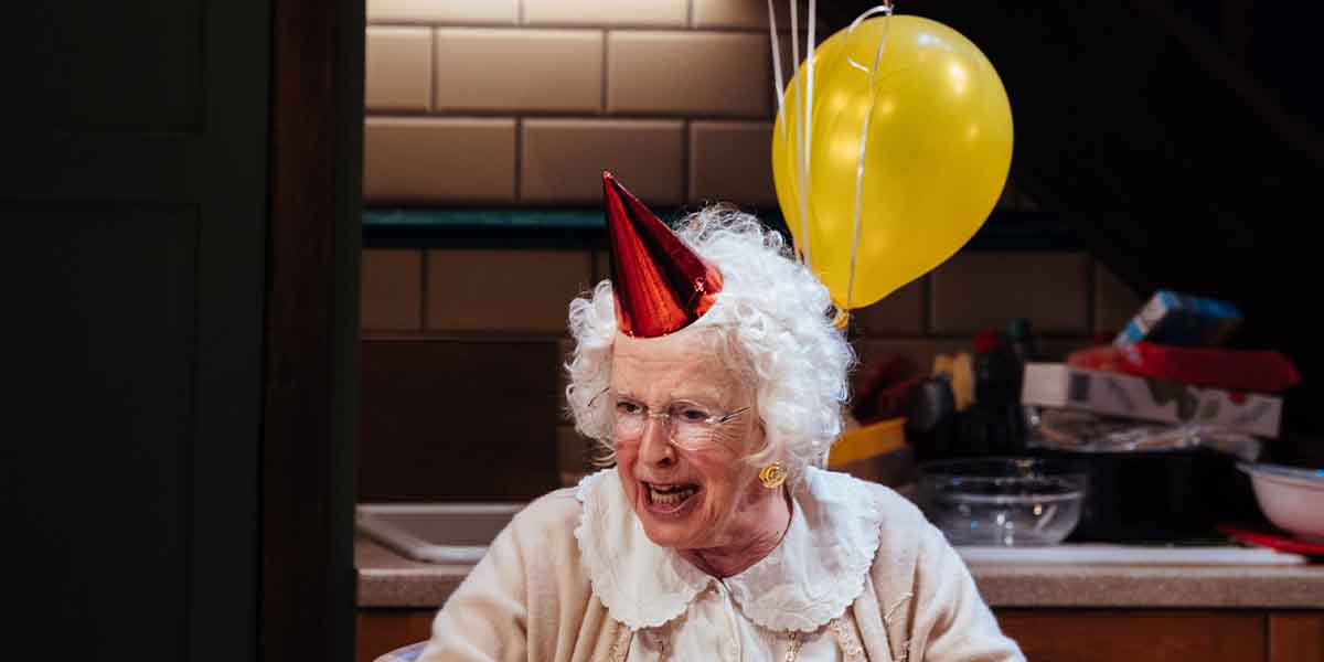 Julia Dearden, wearing a red party hat, smiles animatedly with a yellow balloon in the background.