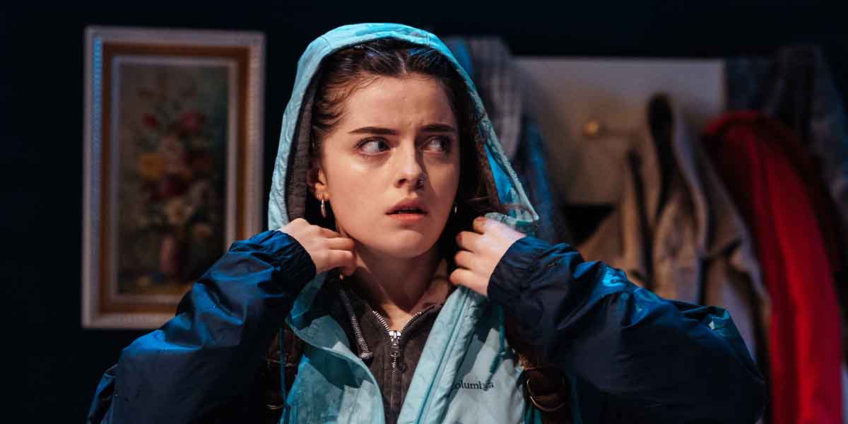 Muireann Ní Fhaogáin looks concerned, adjusting a hooded jacket in a dimly lit room with coats.