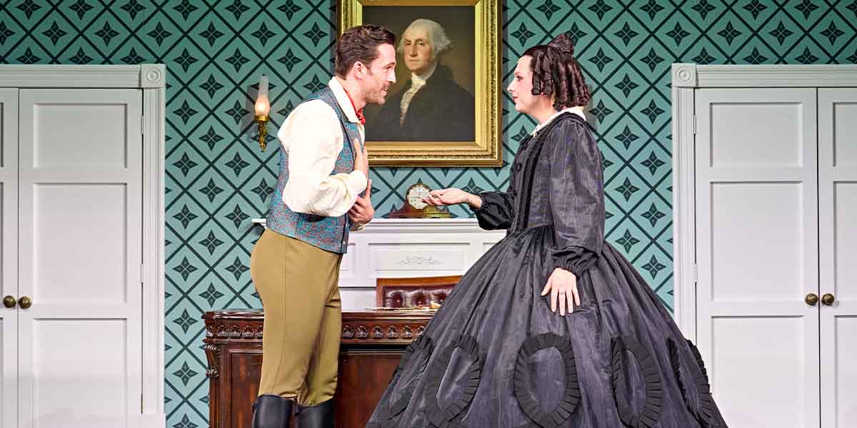A man in period costume converses with a woman in a black dress, against a patterned wallpaper backdrop.