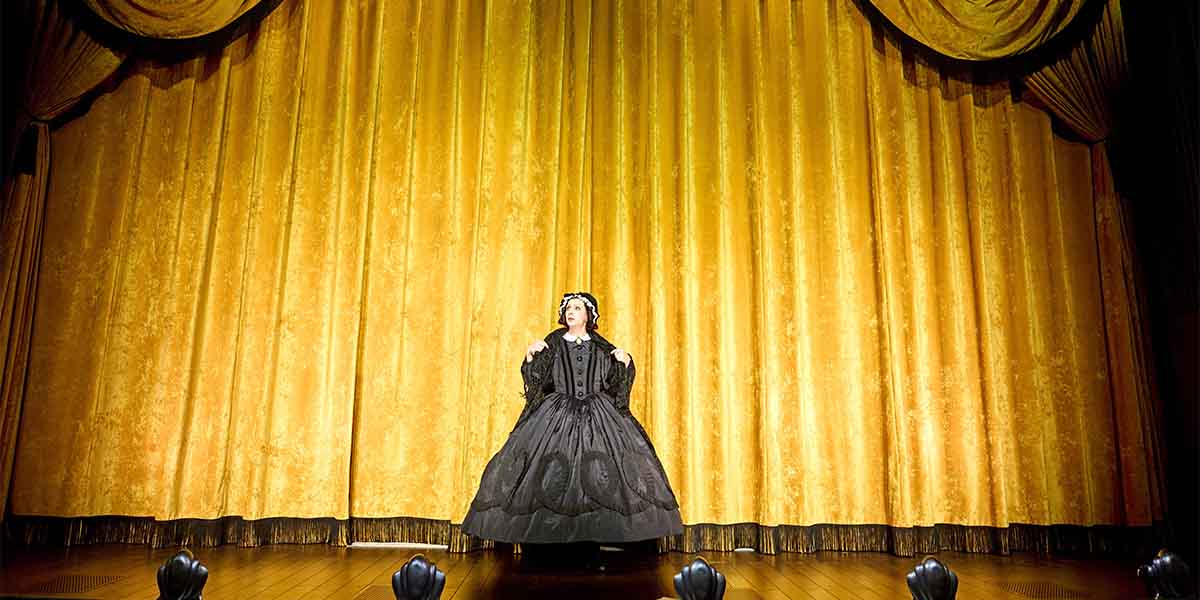 A performer in a black dress stands centre stage, facing an ornate golden curtain backdrop.