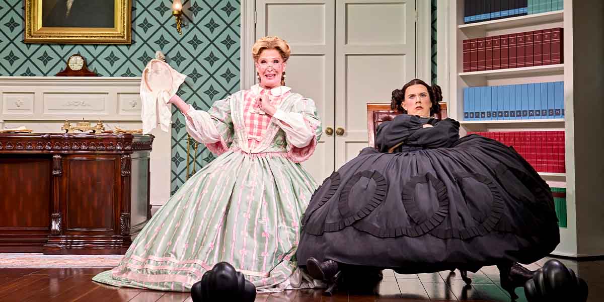 Bianca Leigh and Cole Escola perform on stage, wearing elaborate period costumes, with a wooden desk behind them.