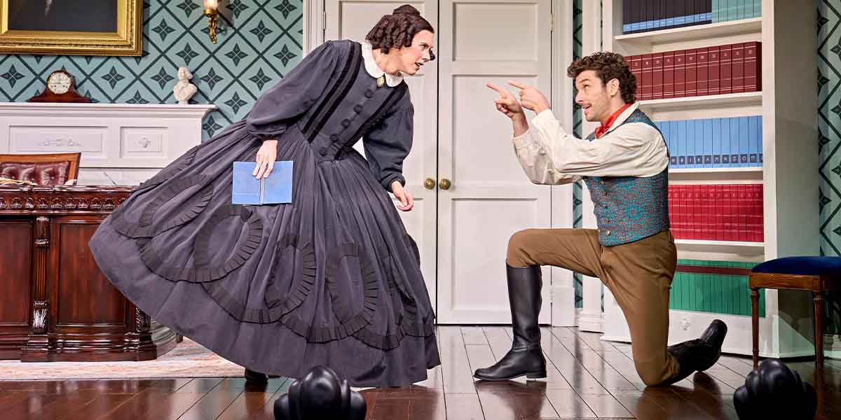 Cole Escola, as Mary Todd Lincoln, gestures with a book, while Michael Urie kneels, animatedly speaking.