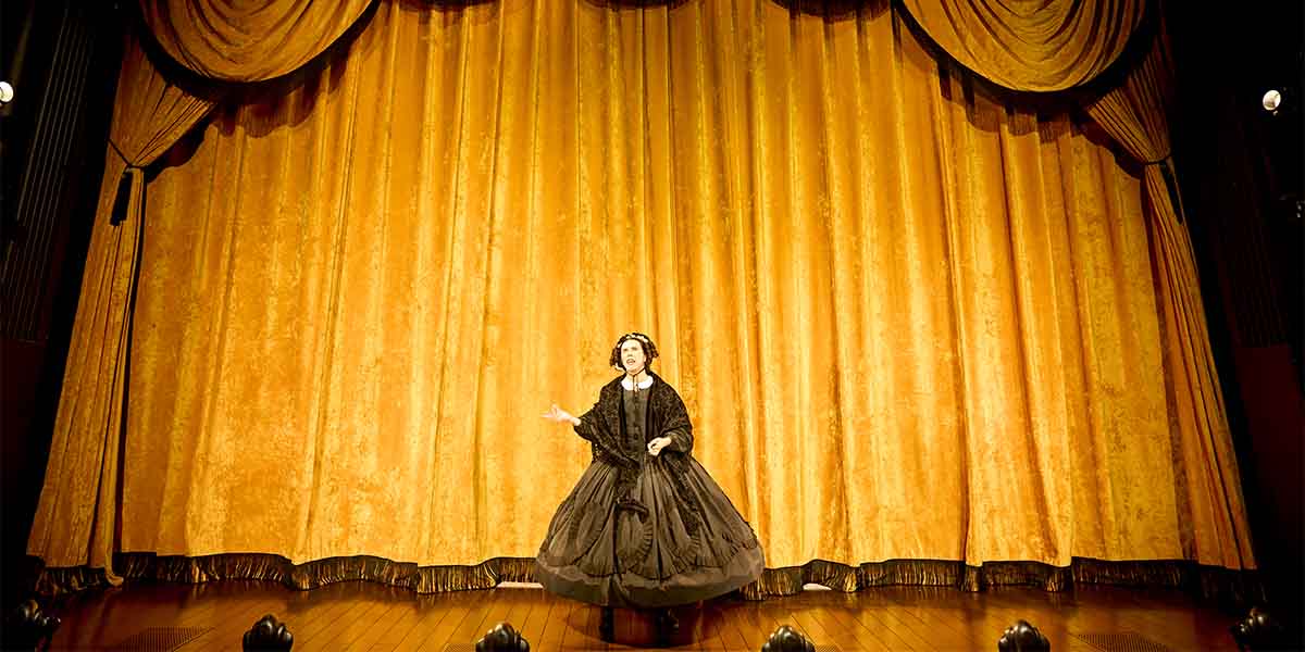 Cole Escola performs as Mary Todd Lincoln on stage, wearing a black dress against a golden curtain backdrop.