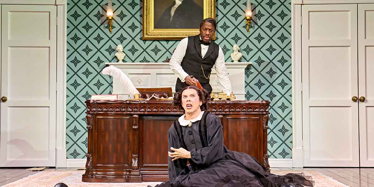 Giles Terera stands at a desk while Cole Escola, in a black dress, kneels in distress.