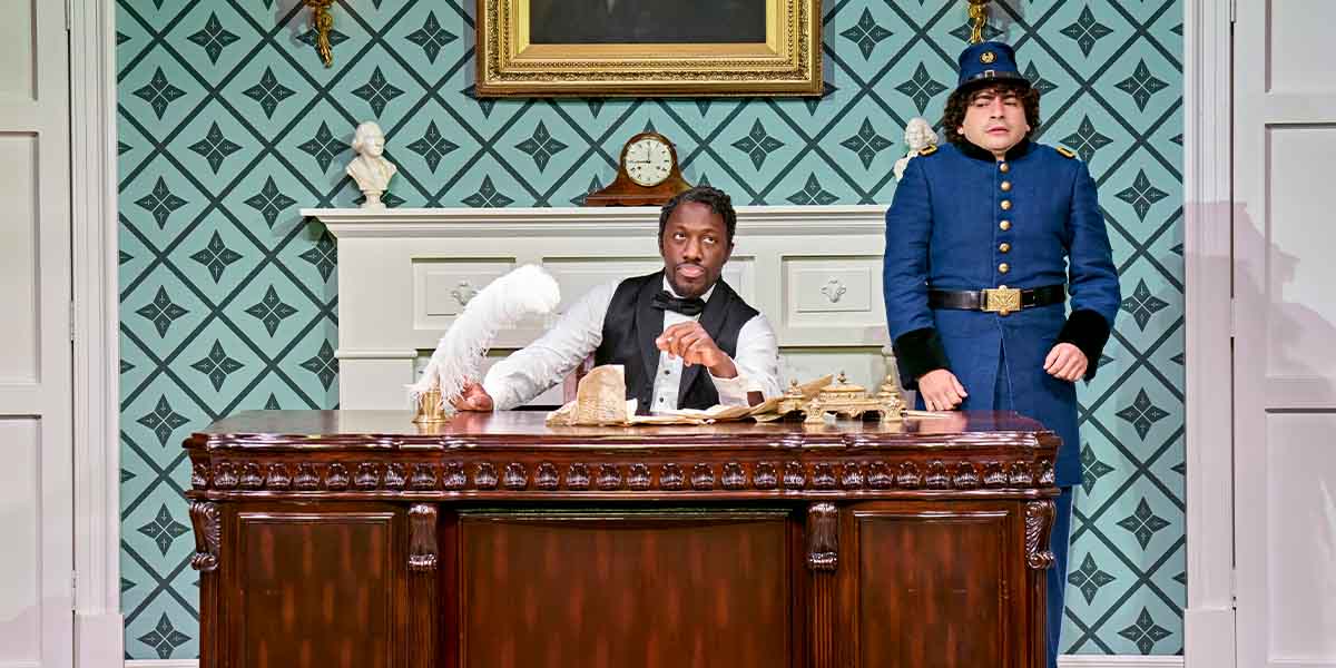 Giles Terera sits at a wooden desk, while Tony Macht stands beside him, both in period costumes.