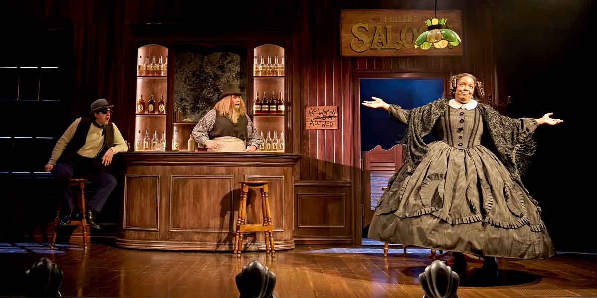 Lucy Frederick, in a dark dress, gestures while standing in a saloon with two men behind the bar.