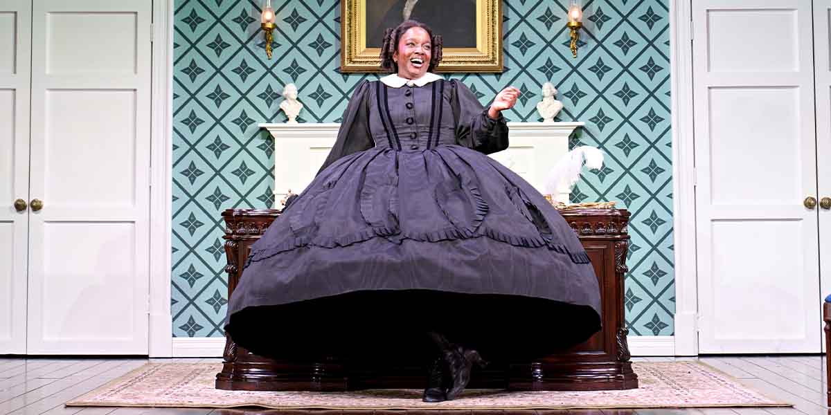 Lolly Adefope, dressed in a large black gown, smiles while gesturing in a vintage room setting.