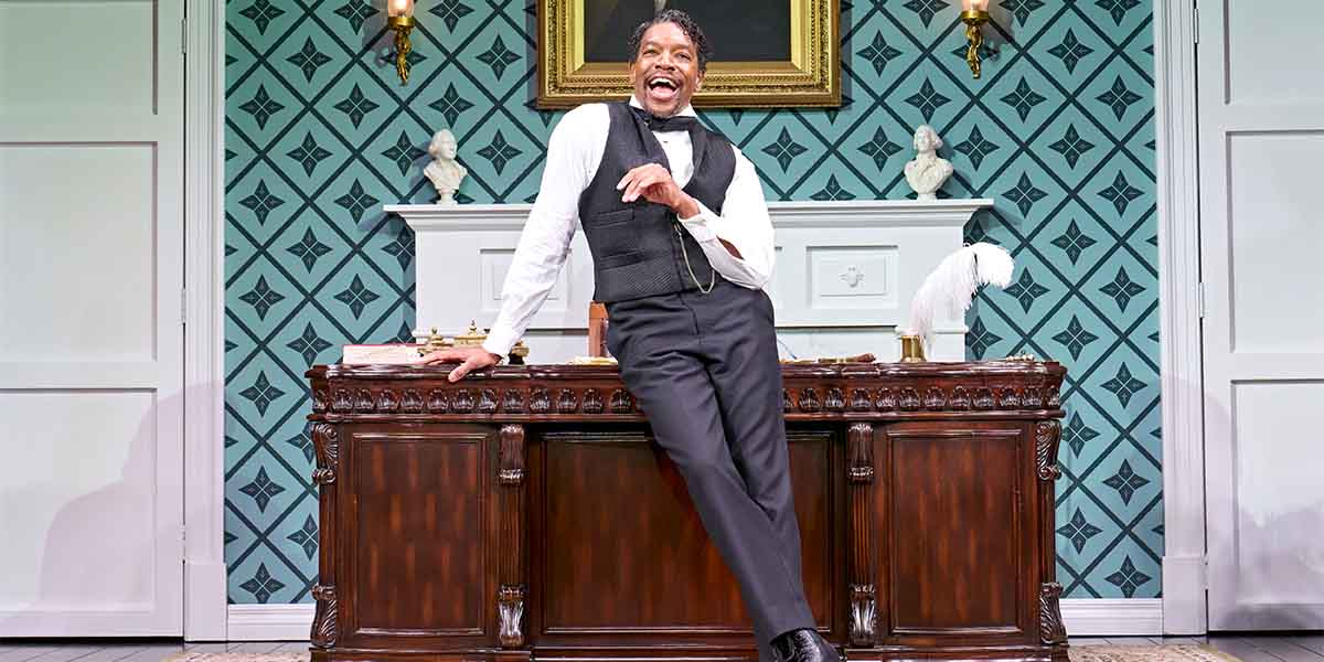 Syrus Lowe, dressed in formal attire, leans playfully on a wooden desk against a patterned wall.