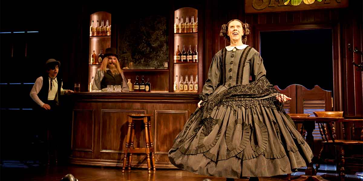 Bianca Leigh, dressed in a black gown, performs energetically on stage with a bar and two men behind.
