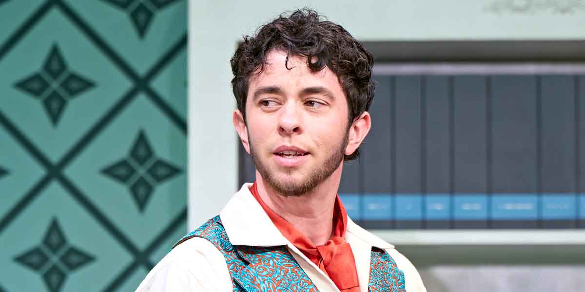 Vinnie Heaven, wearing a patterned vest and red tie, looks thoughtfully to the side against a patterned backdrop.